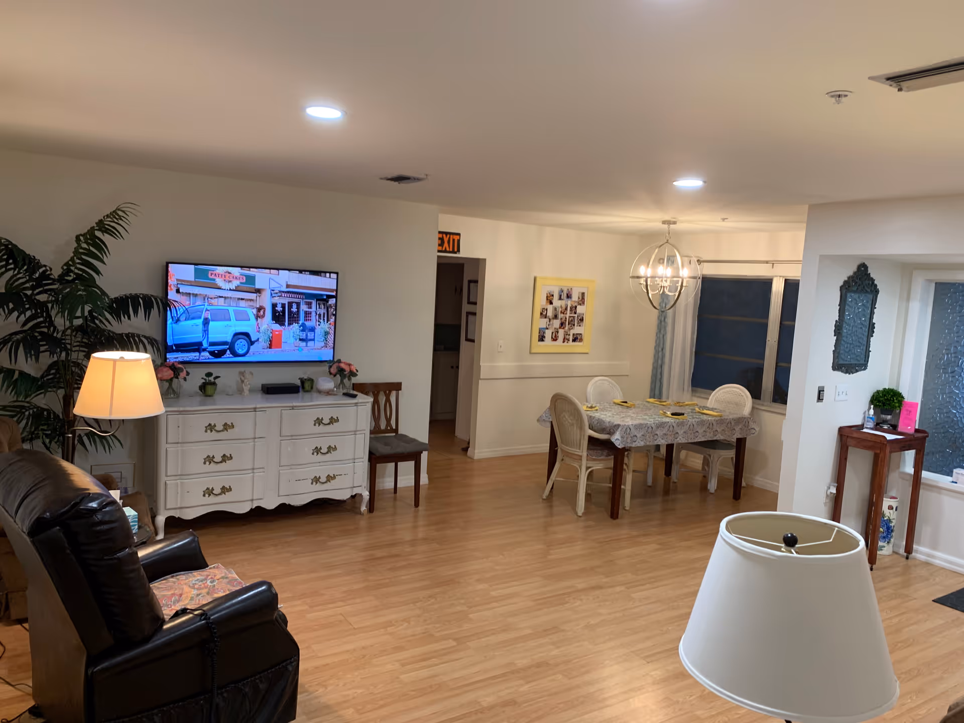 A cozy living and dining area in an assisted living facility with wooden flooring. The living area has a leather recliner chair facing a white dresser with a flat-screen TV mounted above it. There are decorative plants and flowers on the dresser. The dining area features a table covered with a patterned tablecloth and four chairs, set with plates and napkins. The room is well-lit with ceiling lights and a modern chandelier above the dining table. A small wooden side table with a mirror above it is near the window with frosted glass panels.