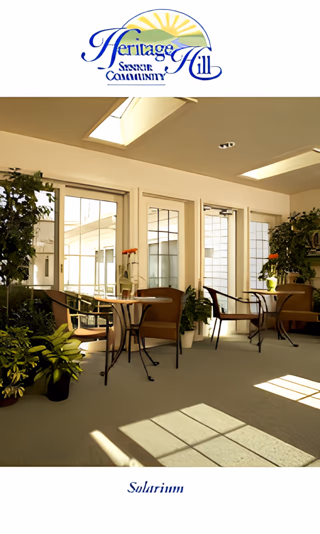 A bright solarium room with large windows and glass doors allowing natural light to flood in. The room contains several small tables with chairs arranged around them, and various green plants placed throughout the space. The ceiling has skylights, and the floor is carpeted. The image includes the Heritage Hill Senior Community logo at the top and the word 'Solarium' at the bottom.