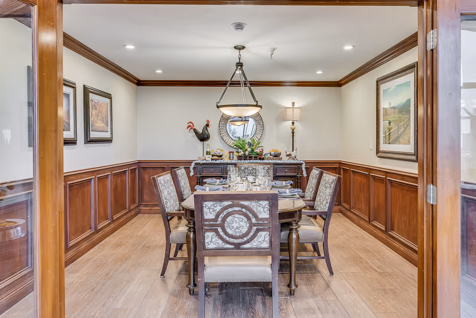A formal dining room with a wooden dining table set for six people. The chairs have patterned upholstery and the room features wood paneling on the lower walls and cream-colored paint above. A sideboard against the back wall holds decorative items including a large rooster figurine, a round mirror, a lamp, and a floral arrangement. The room is illuminated by recessed ceiling lights and a hanging light fixture above the table.