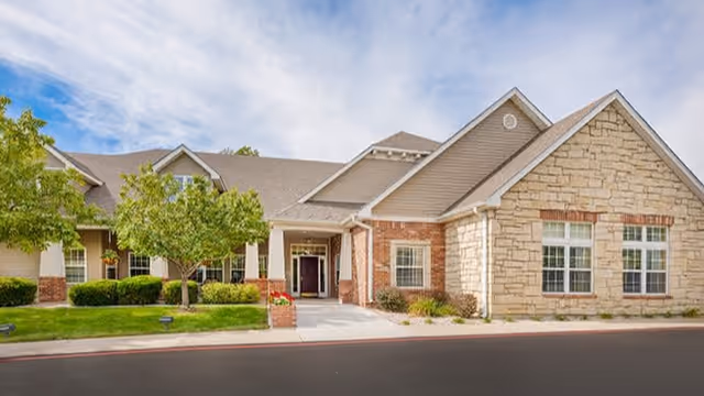 Exterior view of a single-story senior living facility building with a combination of brick and stone facade, multiple windows, a covered entrance, and landscaped greenery including trees and bushes under a partly cloudy sky.