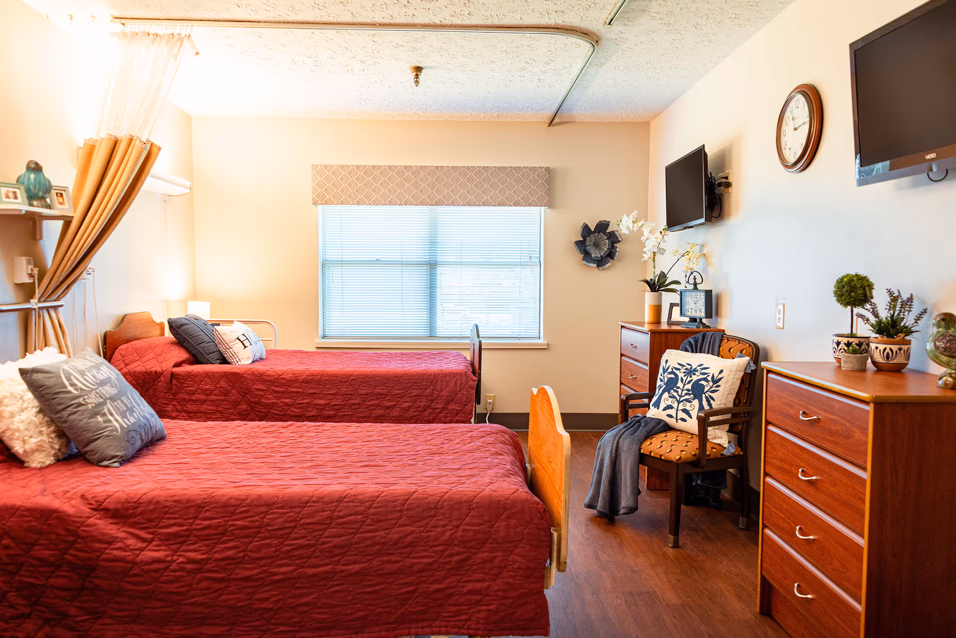 Two twin beds with red bedspreads in a bright, well-decorated senior living bedroom with dressers, a chair, and a wall-mounted TV.