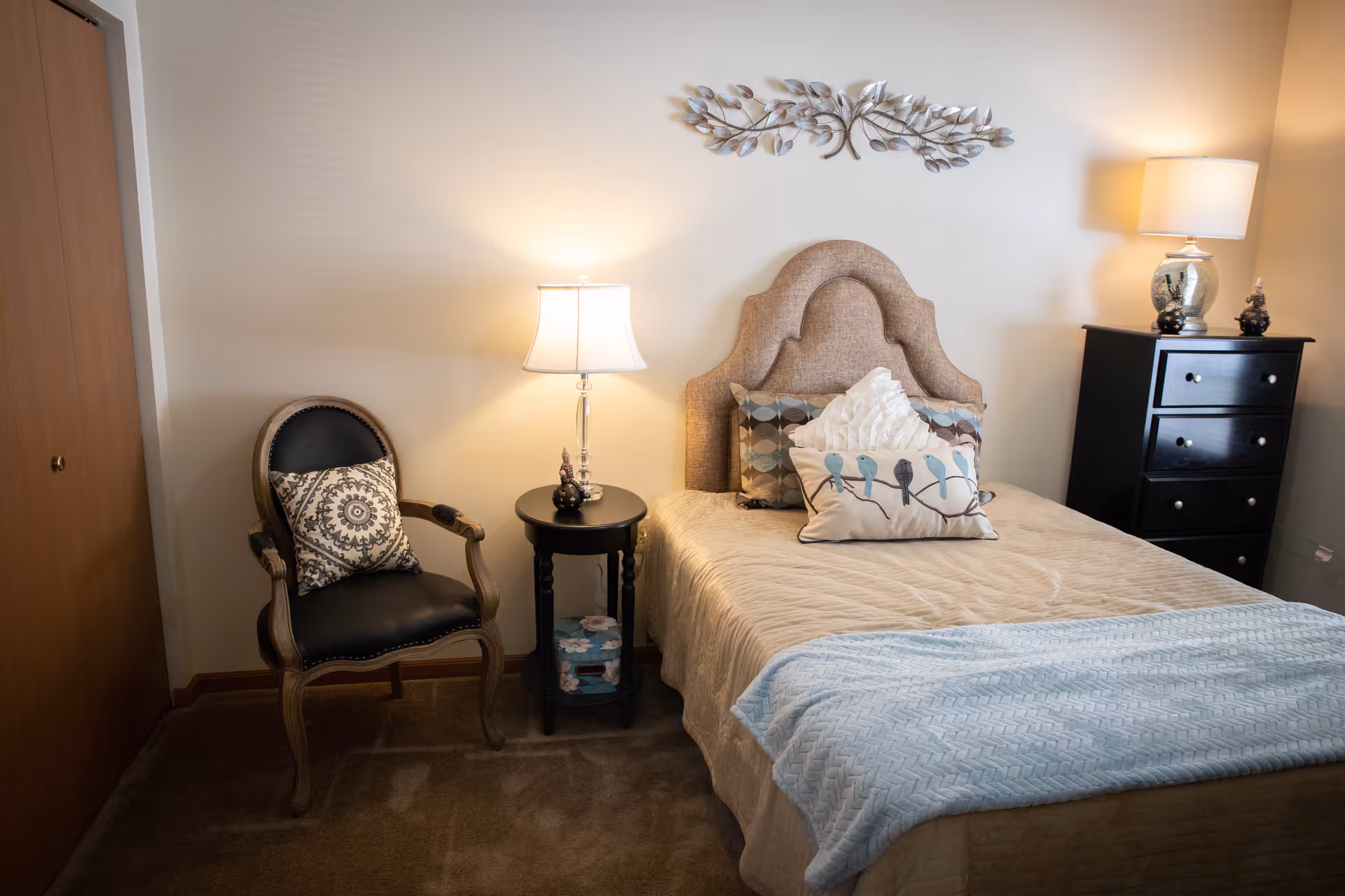A cozy bedroom with a beige upholstered headboard bed, decorated with multiple pillows including one with a bird design. There is a black wooden nightstand with a lamp and decorative items on the left side of the bed, and a black chest of drawers with a lamp on the right side. A wooden chair with a patterned cushion is placed next to the nightstand. The walls are light-colored and there is a decorative metal wall art above the bed.
