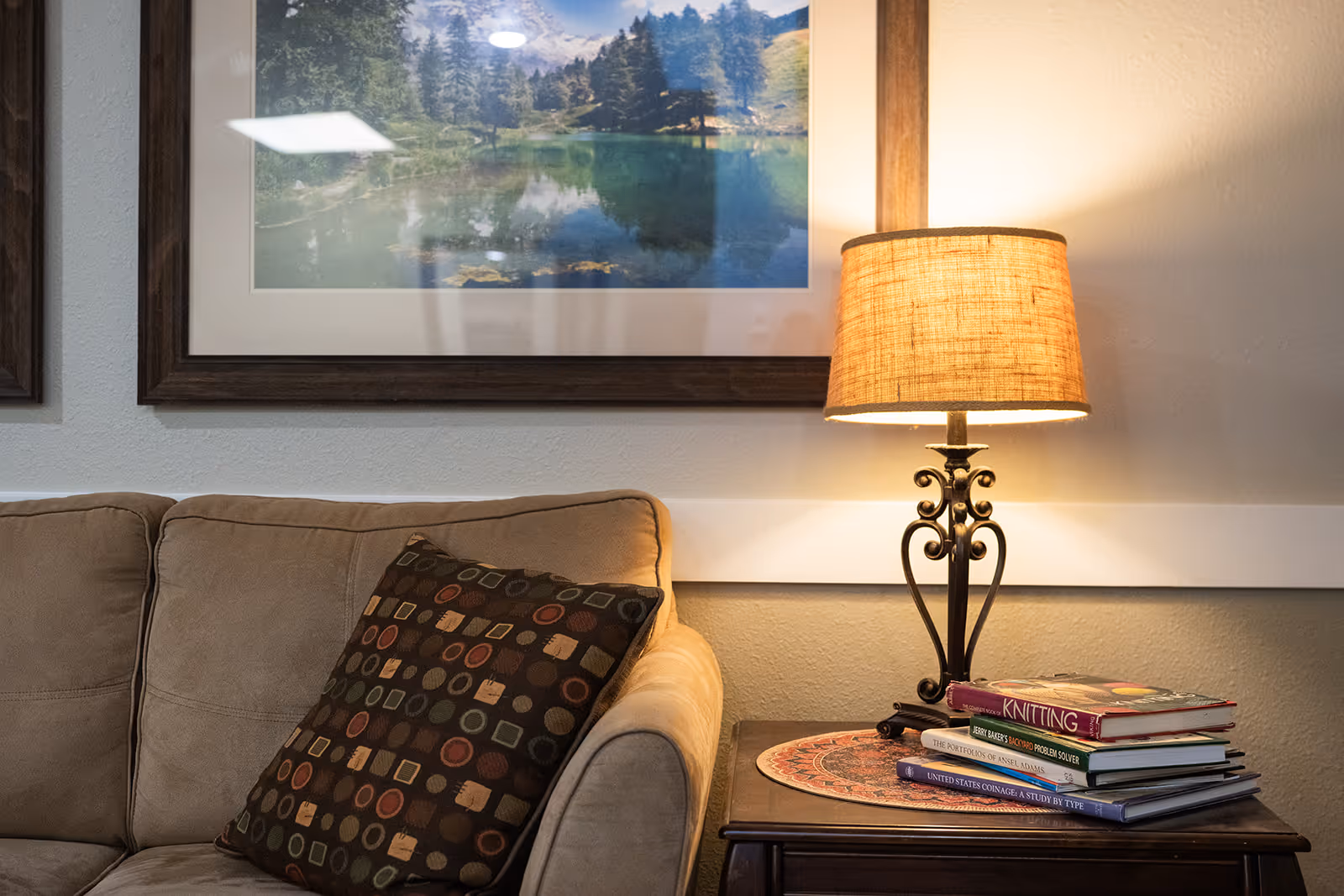 A cozy living room corner with a beige couch featuring a patterned cushion, a wooden side table with a decorative lamp, and a stack of books including one titled 'KNITTING'. Above the couch hangs a framed landscape photograph of a lake surrounded by trees and mountains.
