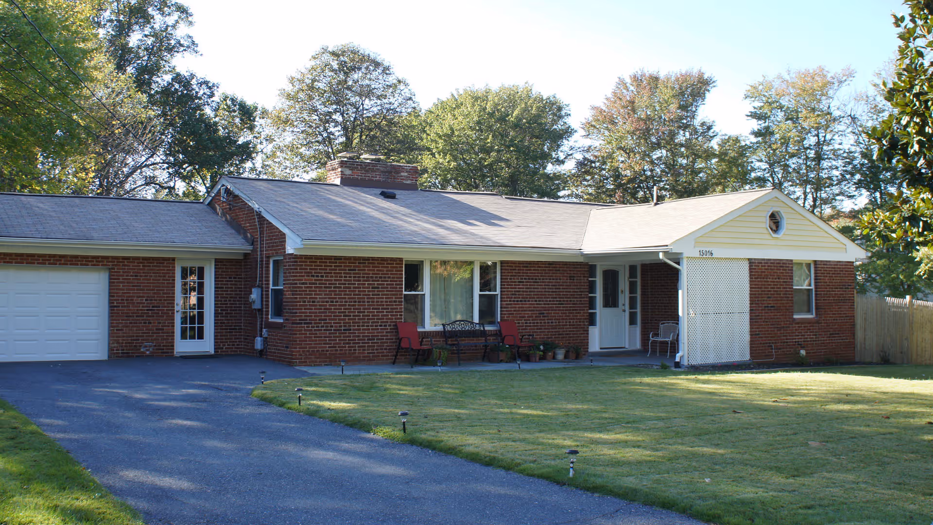 Single-story brick building with a gray shingled roof, a driveway leading to a white garage door on the left, a front porch with chairs and potted plants, and a well-maintained lawn surrounded by trees in the background.