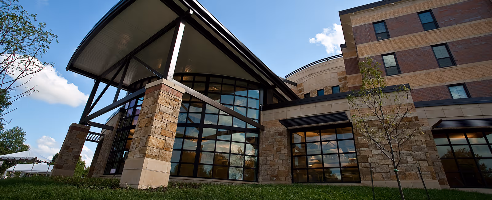 Exterior view of a modern multi-story building with large glass windows, stone and brick facade, and a covered entrance supported by stone pillars under a blue sky with some clouds.