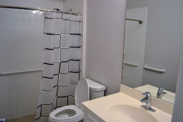 A bathroom with a white toilet with the lid open, a white sink with a silver faucet, a large mirror above the sink, and a shower area with white tiled walls and a white shower curtain featuring a black square pattern. There are white grab bars installed on the tiled walls near the shower and toilet.
