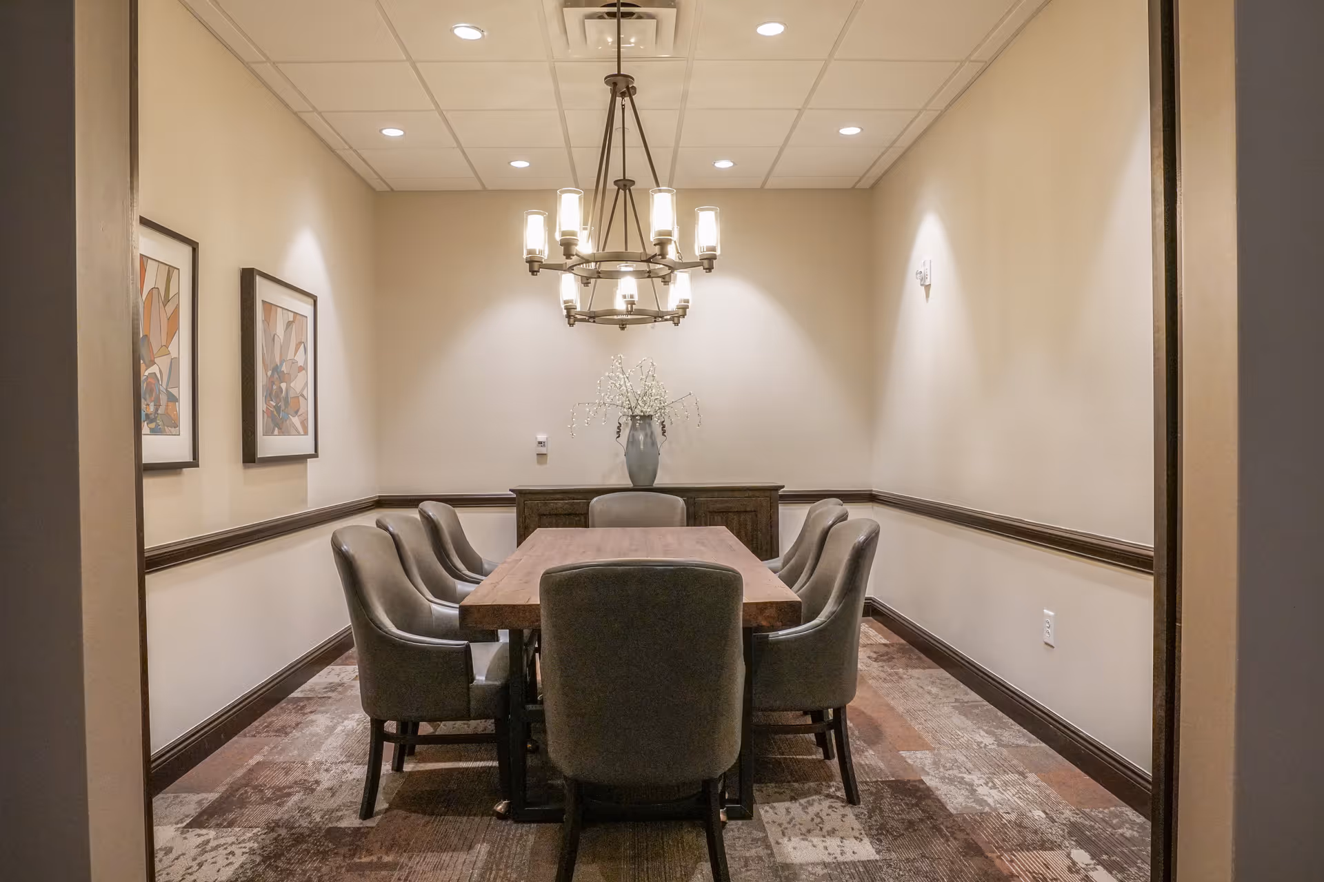 A small, well-lit dining room with a rectangular wooden table surrounded by six cushioned chairs. A modern chandelier hangs above the table, and two framed abstract artworks are on the left wall. At the far end of the room, there is a sideboard with a vase containing decorative branches.