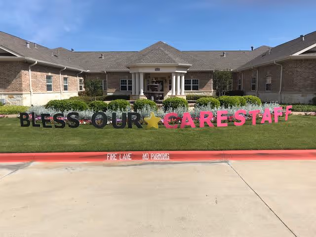 Front entrance of a brick senior living facility with manicured landscaping and large lawn letters spelling 'BLESS OUR CARE STAFF'.