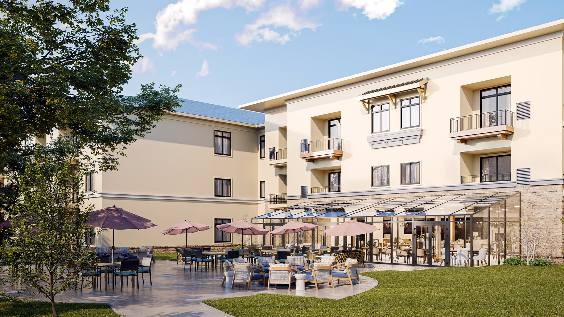 Outdoor patio area of a senior living facility with multiple tables and chairs under purple umbrellas, adjacent to a three-story beige building with balconies and large windows. The patio is surrounded by green grass and trees under a partly cloudy sky.