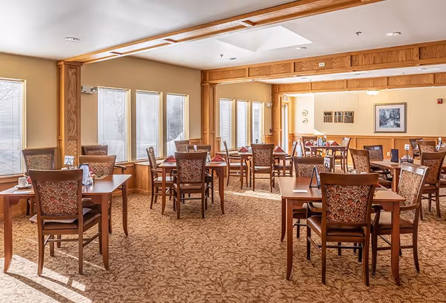 A bright dining room with multiple wooden tables and chairs arranged neatly. Each table has folded red napkins and place settings. Large windows with blinds allow natural light to fill the room. The walls are painted beige with wooden trim, and framed artwork is visible on the far wall.