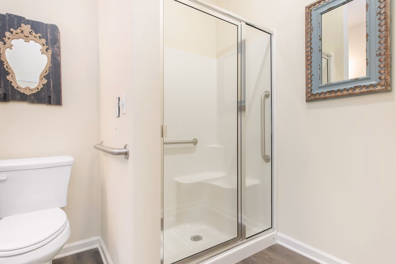 A bathroom with a white toilet, a glass-enclosed shower with a built-in seat and grab bars, and two decorative mirrors on the walls. The walls are painted light beige and the floor has wood-like flooring.