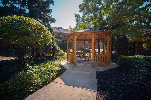 A wooden gazebo with chairs inside situated in a garden area surrounded by trees and shrubs, with a brick building visible in the background under a clear sky.