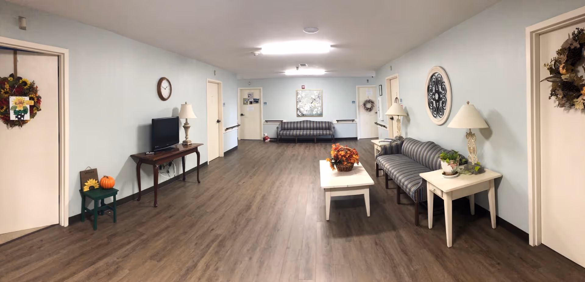 A spacious common area in an assisted living facility with light blue walls and wood flooring. The room features two striped sofas, a white coffee table with a basket of autumn leaves, two white side tables with lamps and plants, a wall clock, a flat-screen TV on a wooden table, and several doors decorated with wreaths. The ceiling has fluorescent lighting.