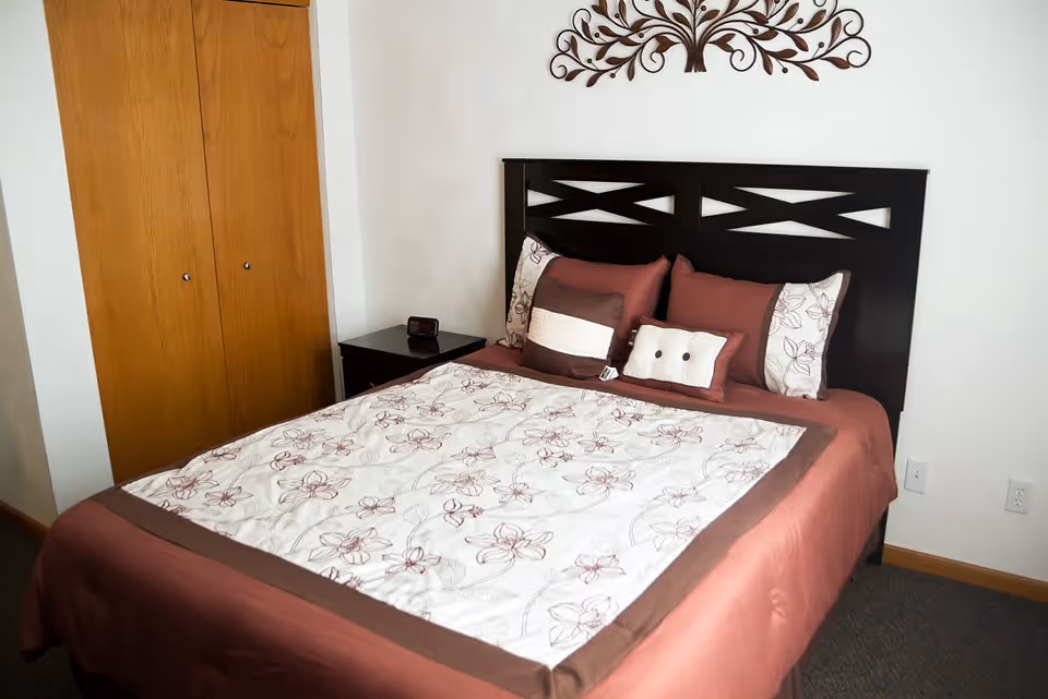 A bedroom with a neatly made bed featuring a floral patterned white and brown bedspread and matching pillows. The bed has a dark wooden headboard. To the left of the bed is a small dark wooden nightstand with a digital alarm clock on it. There is a wooden closet with double doors on the left side of the room. The walls are white and there is a decorative metal wall art piece above the headboard.