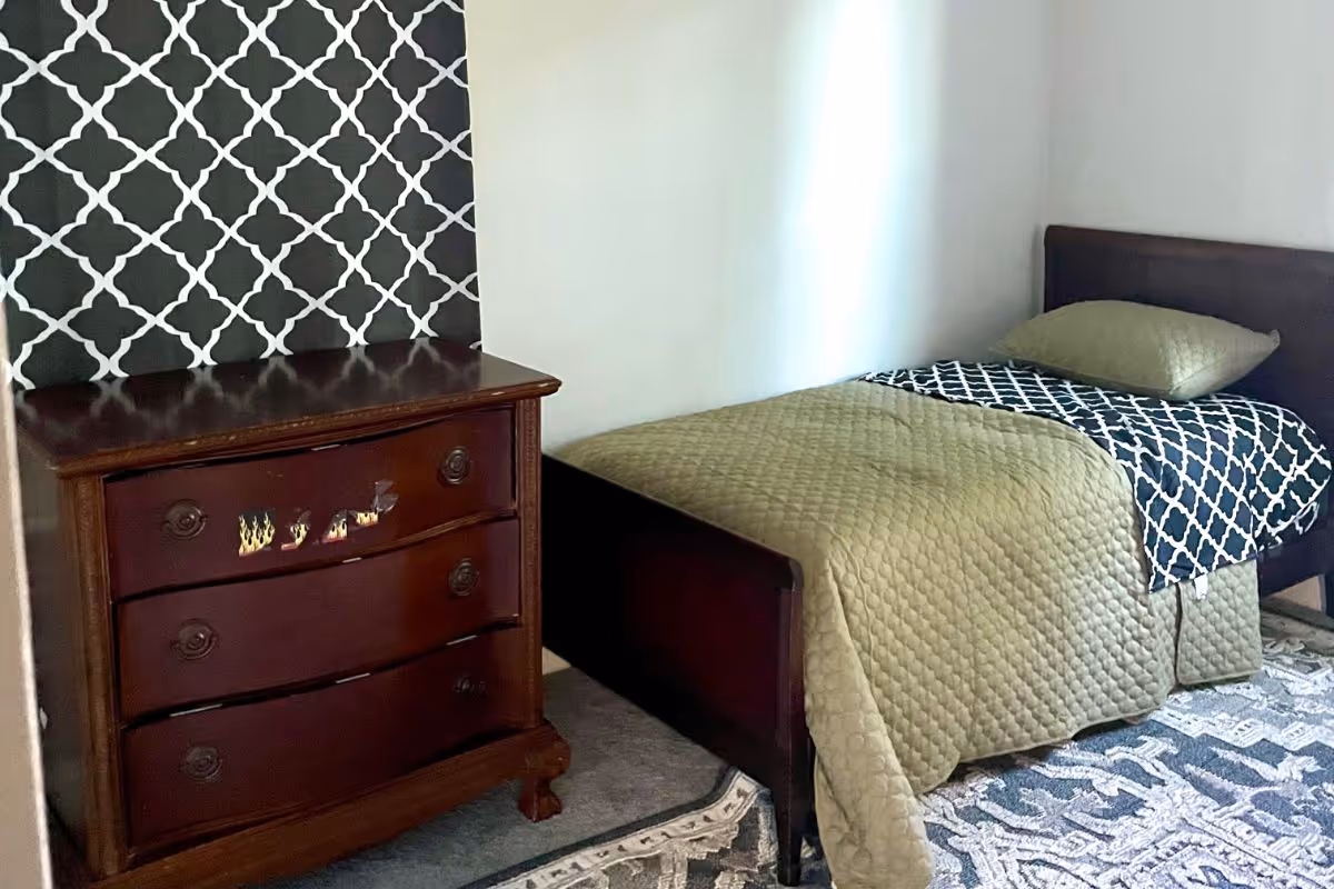 A tidy bedroom with a wooden dresser, bed, patterned wallpaper, and a rug.