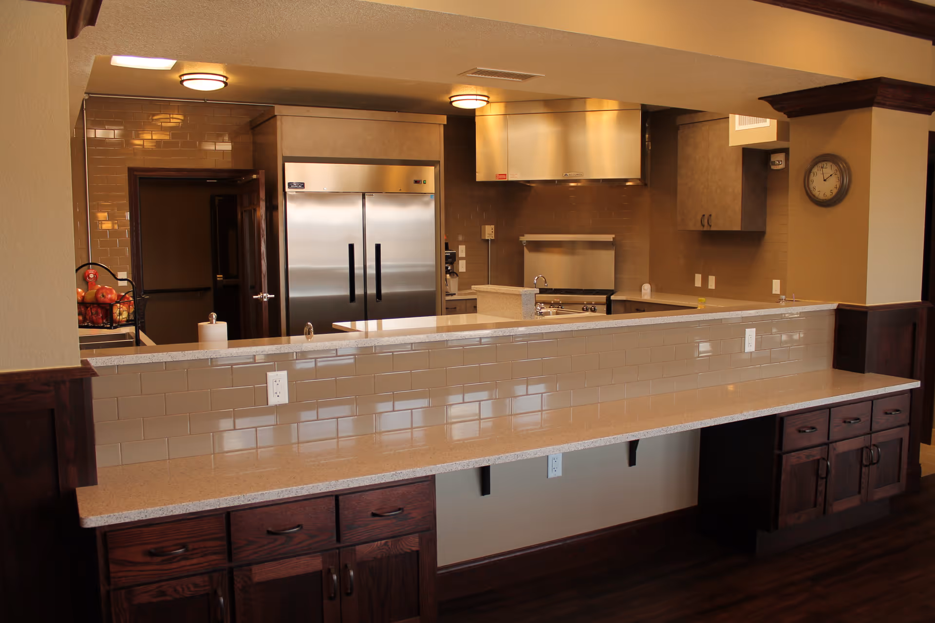Interior view of a modern kitchen area with a long countertop and backsplash tiles. The kitchen features stainless steel appliances including a double-door refrigerator and a range hood. There are wooden cabinets below the counter and wall-mounted cabinets, with a clock on the wall. The lighting is warm and the floor is dark wood.