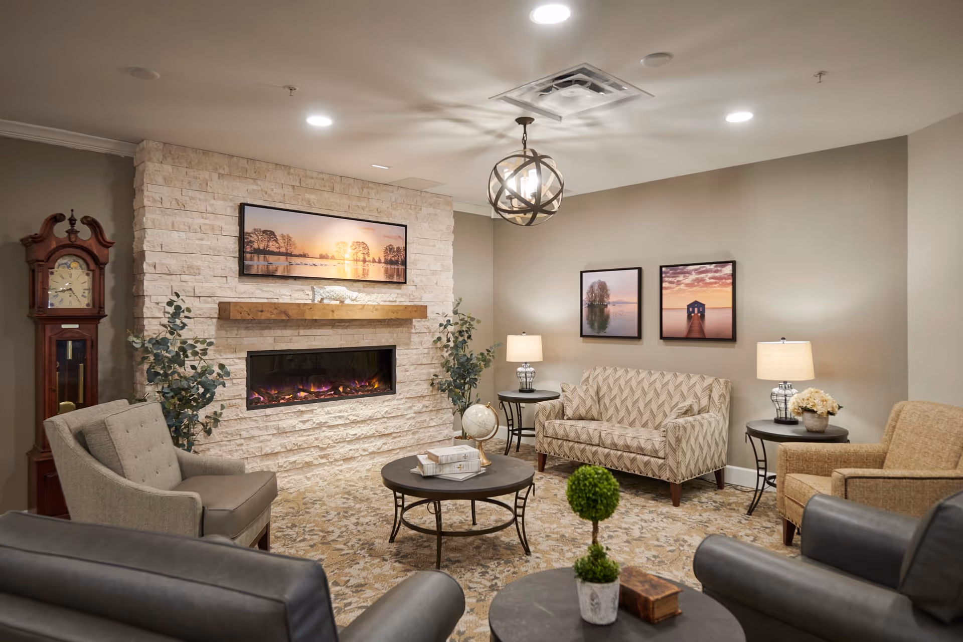 A cozy senior living facility lounge area with a modern electric fireplace set in a stone wall. The room features a grandfather clock, two armchairs, a patterned loveseat, two side tables with lamps, a round coffee table with books, and decorative plants. Three framed landscape photos hang on the walls, and a spherical chandelier hangs from the ceiling.