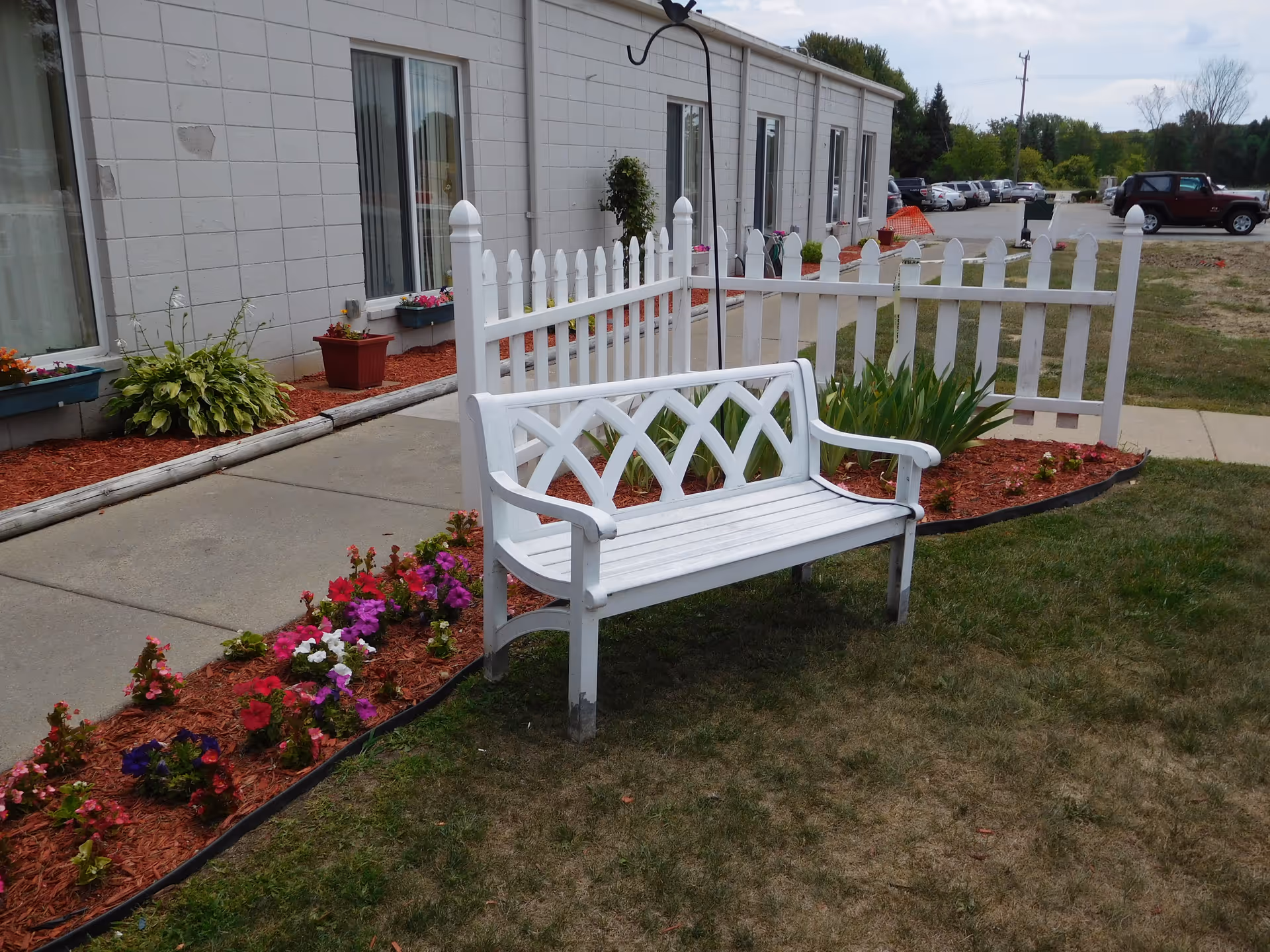 A white wooden bench sits in a landscaped garden with flowers and a white picket fence in front of a single-story building.