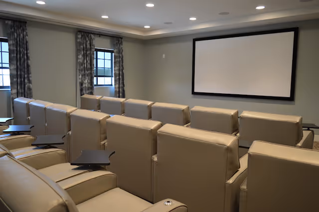 A small theater room with multiple rows of beige leather recliner chairs facing a large blank projection screen on a light gray wall. The room has recessed ceiling lights and two windows with patterned curtains on the left side.