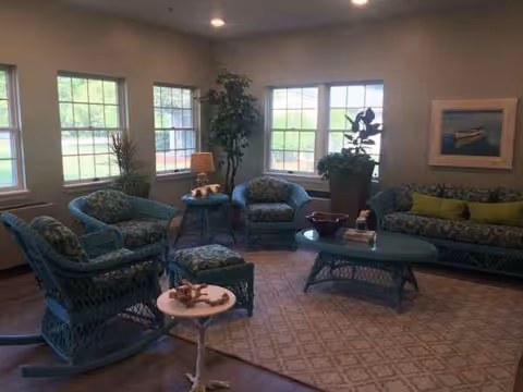 Sunlit senior living lounge with blue wicker seating and potted plants.