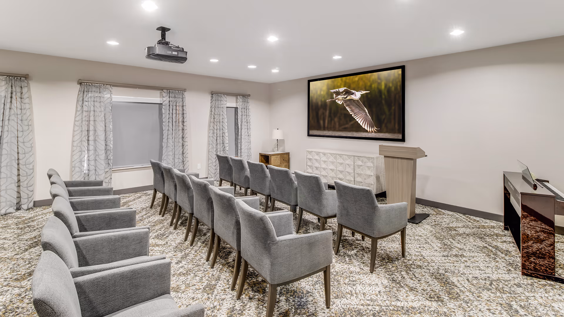 A small conference or presentation room with two rows of gray upholstered chairs facing a podium and a large wall-mounted screen displaying a bird in flight. The room has patterned carpet, light-colored walls, three windows with patterned curtains, a piano on the right side, and a ceiling-mounted projector.