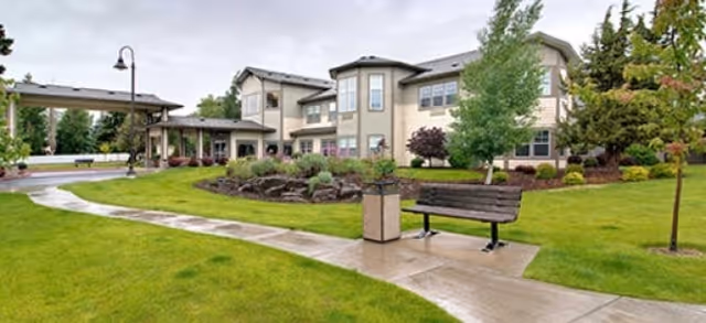Exterior view of a senior living facility with a well-maintained lawn, a curved concrete walkway, a wooden bench, a trash can, and several trees and shrubs under a cloudy sky.