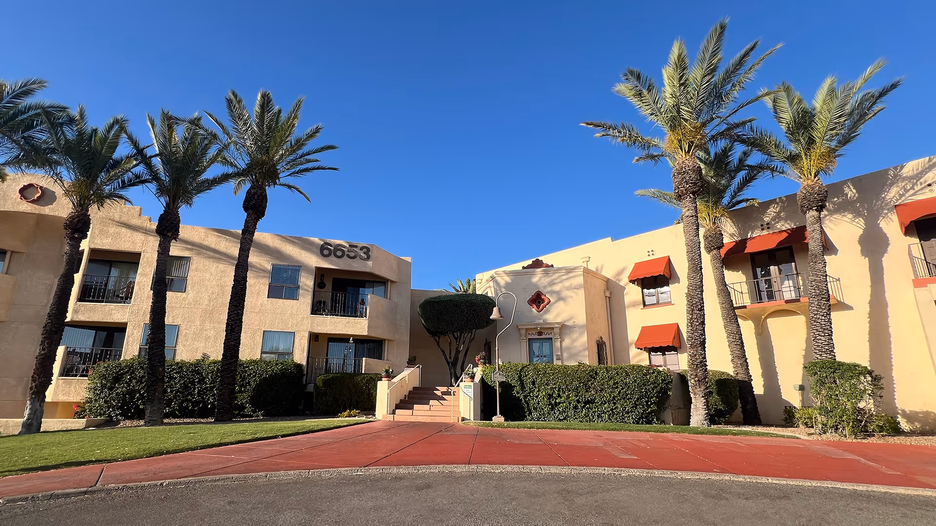 Exterior view of a beige stucco building with the number 6653 displayed prominently. The building has balconies with railings and red awnings over some windows. There are several tall palm trees and neatly trimmed bushes in front of the building under a clear blue sky.