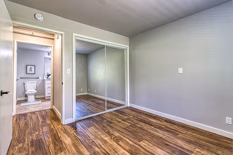 Empty bedroom with wood-look flooring, mirrored sliding closet doors, and a view into an adjoining bathroom.