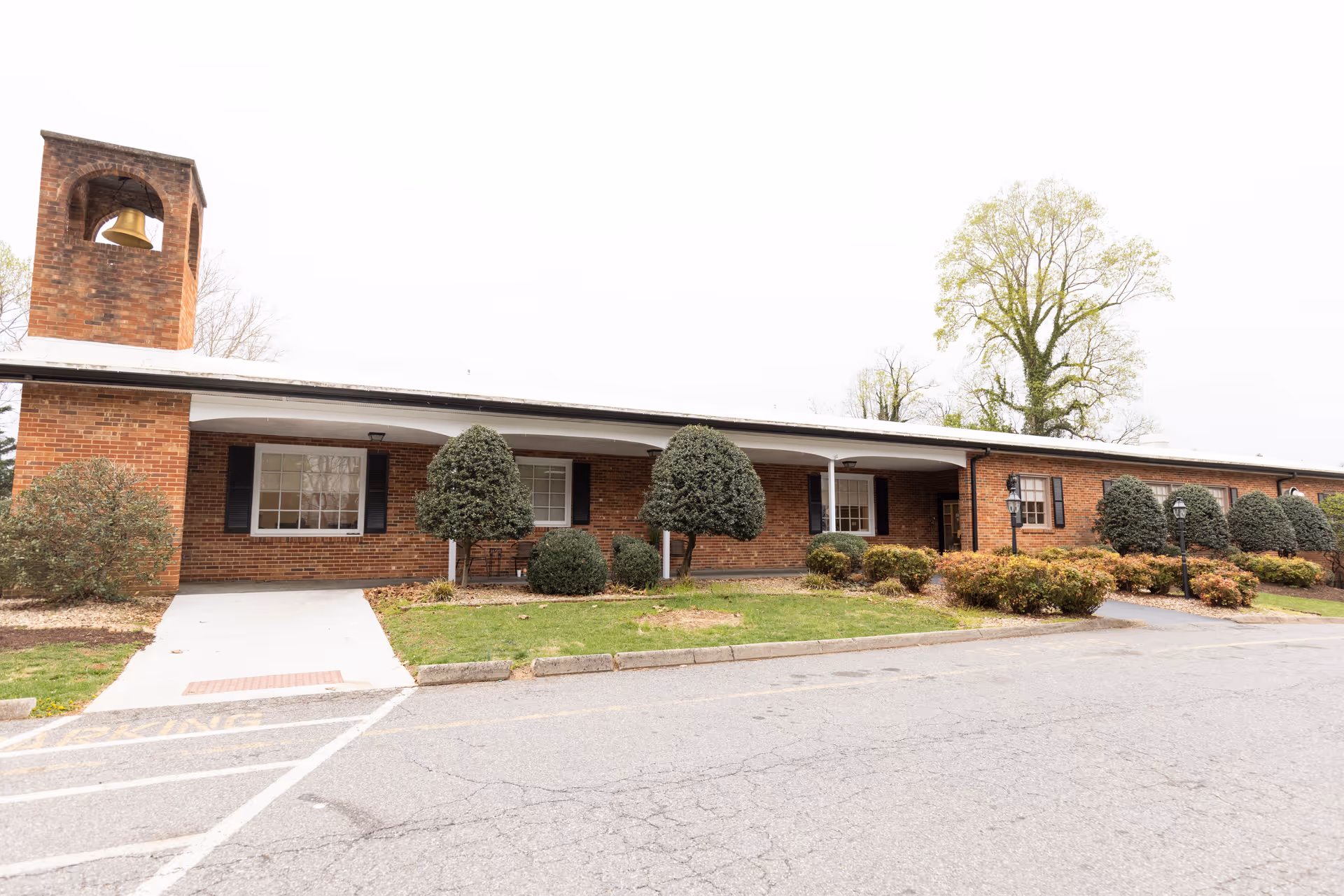 Exterior view of a single-story brick building with a bell tower on the left side. The building has several windows with black shutters and a covered walkway supported by white columns. There are neatly trimmed bushes and small trees in front of the building, along with a paved driveway and parking area.