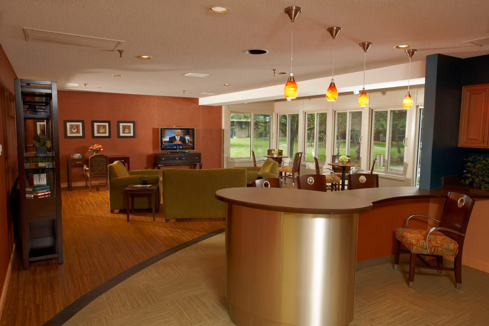 A cozy common area in Canterbury Nursing and Rehabilitation Center featuring a curved reception desk with a chair, pendant lights hanging from the ceiling, green sofas facing a television on a stand, a small desk with a chair and flowers, a tall bookshelf, and several small tables with chairs near large windows overlooking a green outdoor space.