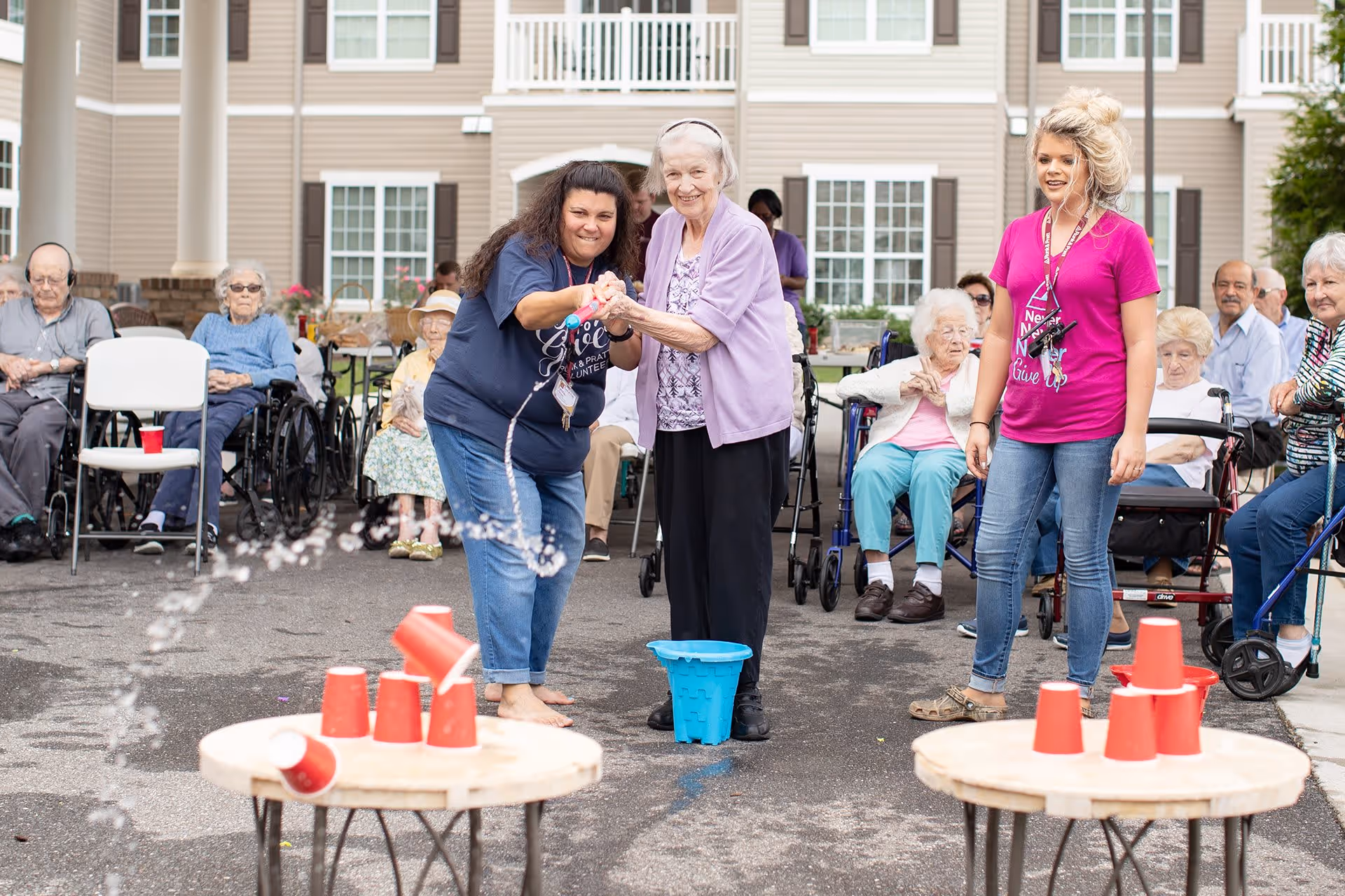 An outdoor scene at a senior living facility where elderly residents and staff are participating in a game involving knocking over red plastic cups stacked on small round tables. Two women, one elderly and one staff member, are actively engaged in the game while other residents, some in wheelchairs, watch and enjoy the activity in front of a beige building with white windows and columns.