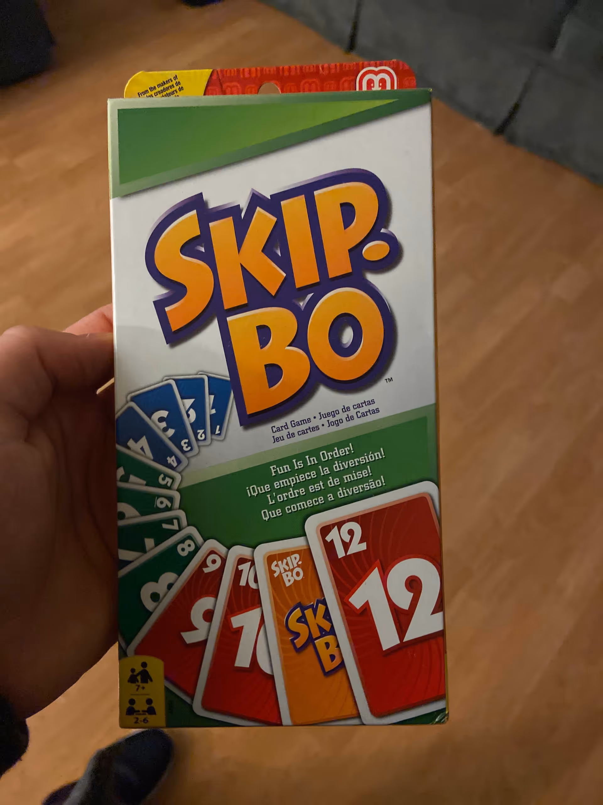 A hand holding a Skip-Bo card game box with a hardwood floor and a couch visible in the background.
