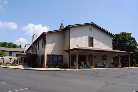 Exterior view of a two-story senior living facility building with beige siding and red trim around the windows. The building has a covered porch area with rocking chairs and some hanging plants. The sky is clear with a few clouds, and there is a paved driveway and parking area in front of the building.