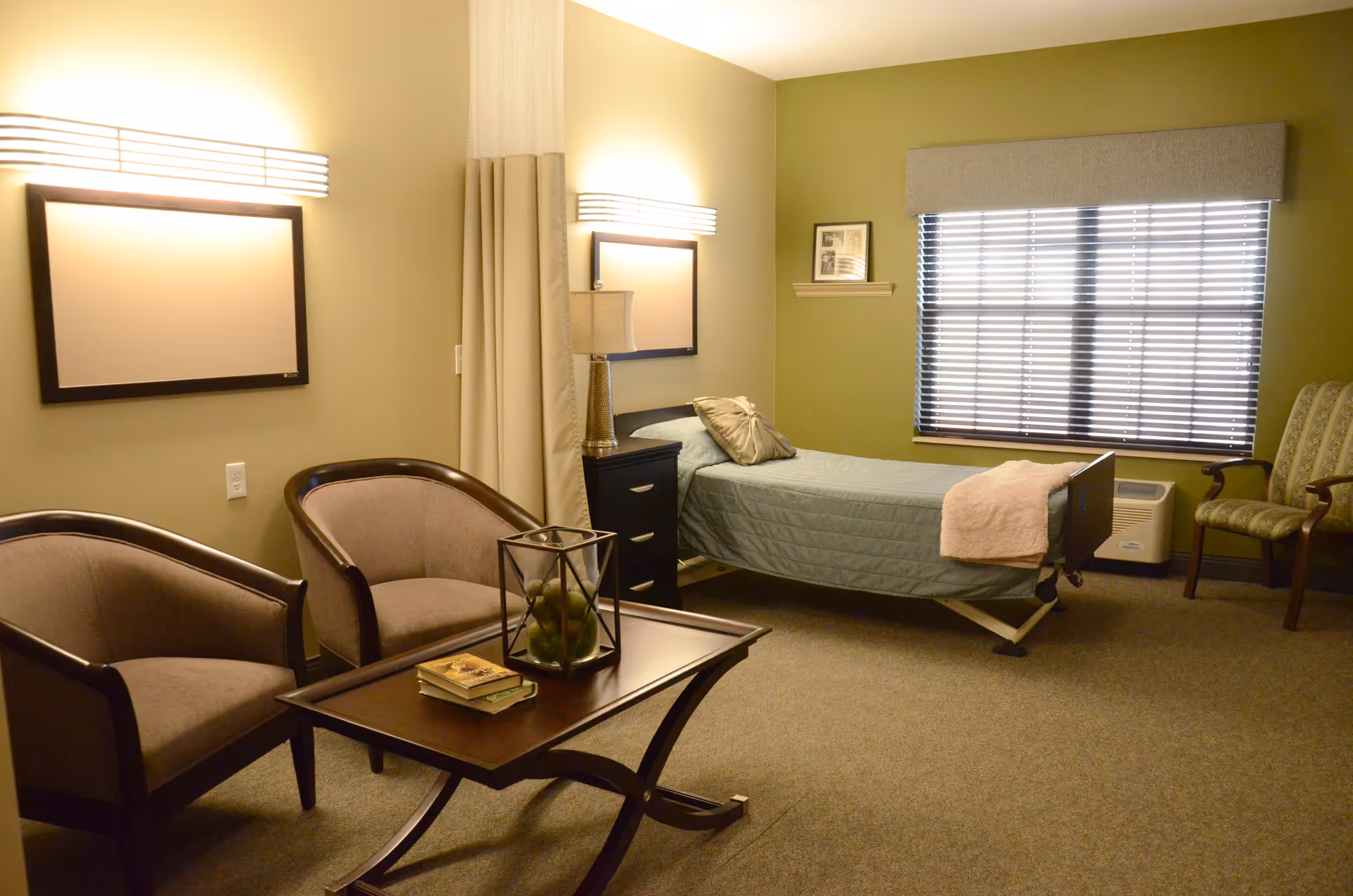 A furnished senior living bedroom with a single adjustable bed, two armchairs, a coffee table and a window with blinds.