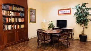 A cozy interior room with a wooden bookshelf filled with books on the left, a round wooden table with four chairs in the center, a small TV on a stand against the wall, a framed picture above the TV, a floor lamp, and a tall potted plant in the corner.