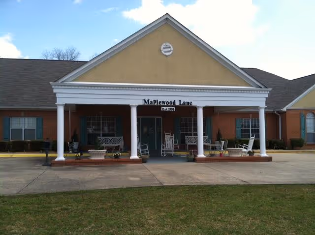 Front exterior view of Maplewood Lane Assisted Living facility showing a single-story brick building with a covered porch supported by white columns. The porch has several white rocking chairs and benches. The building has a triangular pediment with the name 'Maplewood Lane Est. 1993' displayed above the entrance. The sky is partly cloudy.