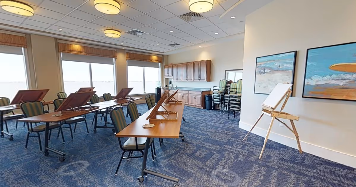 A bright and spacious activity room with large windows letting in natural light. The room contains several tables with adjustable wooden drawing boards and green cushioned chairs on wheels. There is a wooden easel with a blank canvas near two colorful paintings on the wall. In the background, there are wooden cabinets and a stack of additional chairs. The carpet is blue with a patterned design, and the ceiling has recessed lighting and round light fixtures.