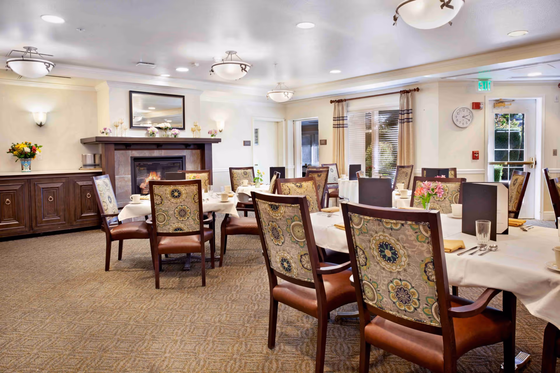 A dining room with several tables covered in white tablecloths, each set with cups, glasses, and napkins. The chairs have patterned backs and leather seats. There is a fireplace with a mirror above it, floral decorations on the mantel, and a sideboard with a vase of flowers. The room has beige walls, carpeted floor, ceiling lights, and windows with curtains letting in natural light.