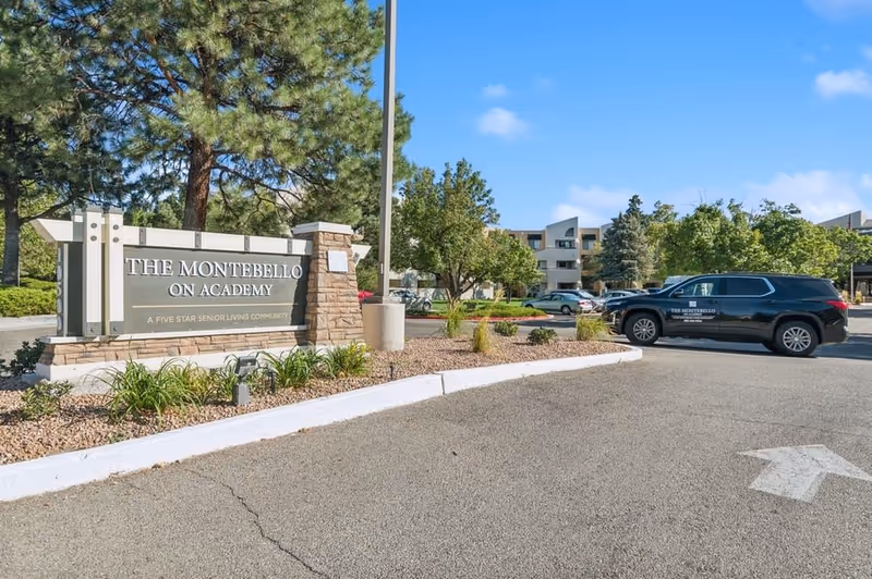Entrance sign for The Montebello on Academy senior living community with landscaping and a parking lot featuring a black vehicle with the facility's logo. Trees and a multi-story building are visible in the background under a clear blue sky.