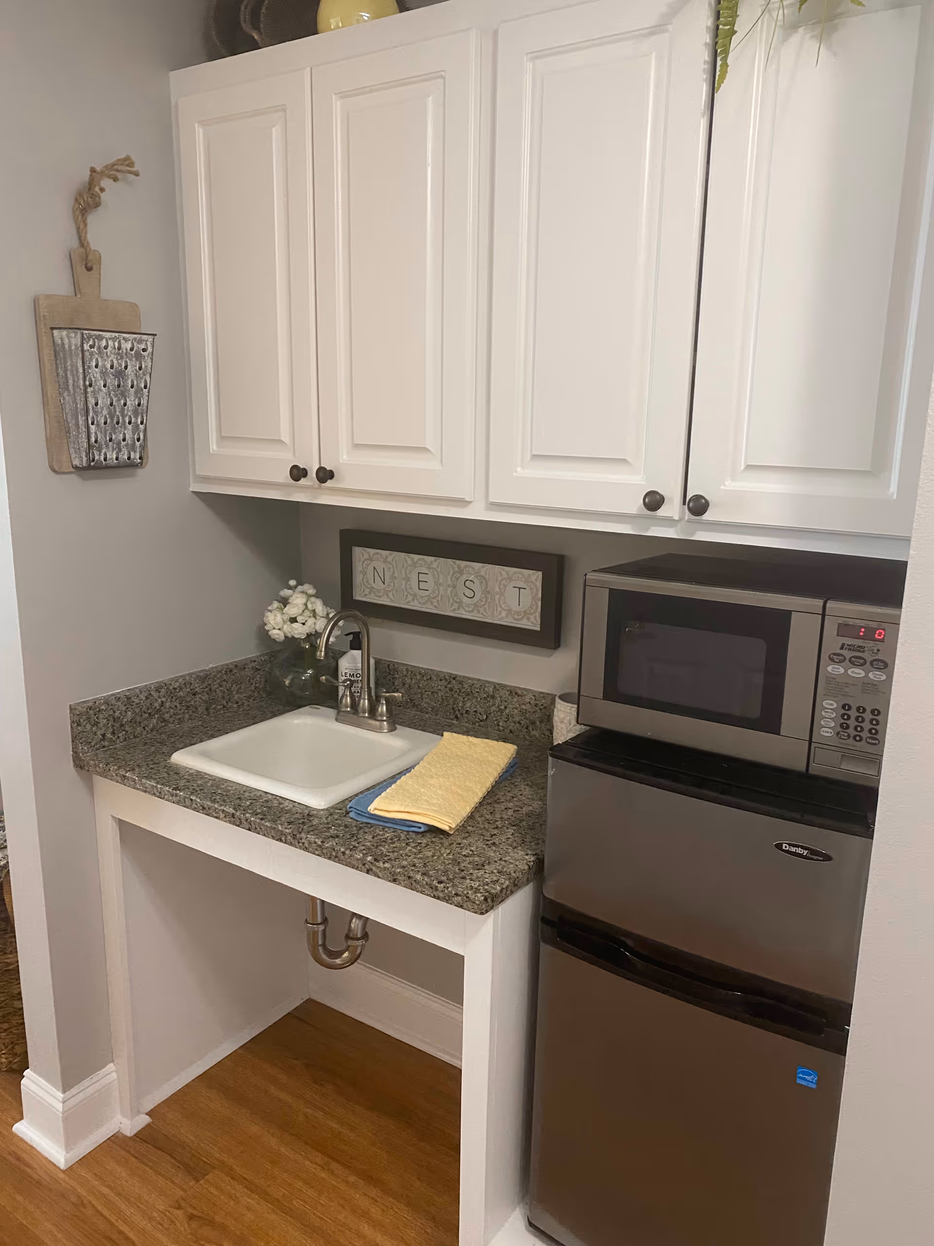 A small kitchenette area with white cabinets above a granite countertop. The countertop has a white sink with a silver faucet, a small glass vase with white flowers, and two folded towels in yellow and blue. To the right of the sink is a stainless steel microwave placed on top of a matching mini refrigerator. The wall behind the sink has a decorative sign that reads 'NEST'. The floor is wooden, and there is a small decorative hanging on the left wall.