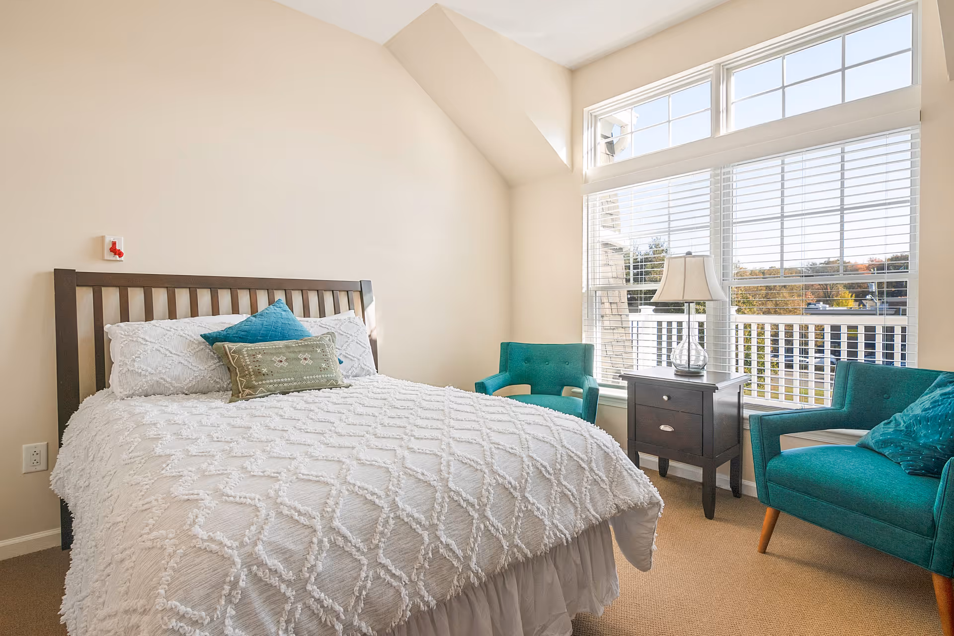 A bright bedroom with a large window letting in natural light. The room features a bed with a white textured comforter and two decorative pillows, a dark wooden nightstand with a lamp, and two teal armchairs positioned near the window. The walls are painted a light beige color.
