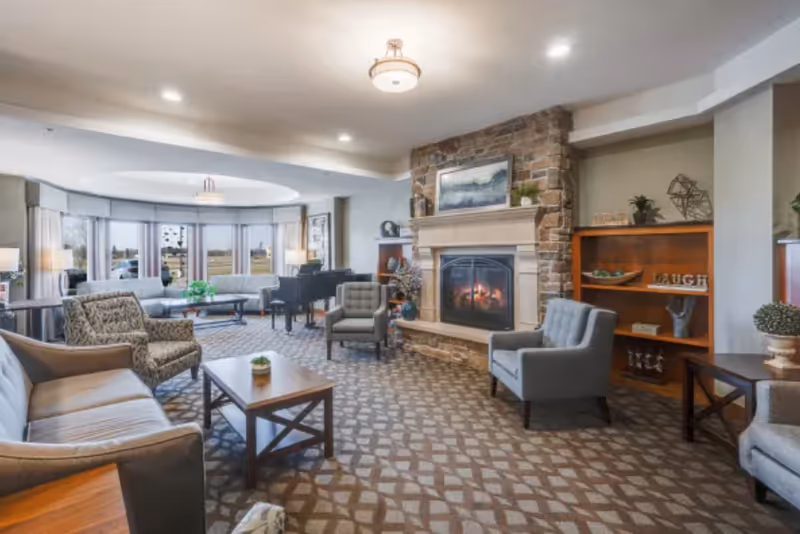 A spacious senior living common area with comfortable seating including sofas and armchairs arranged around a coffee table. There is a stone fireplace with a fire burning and a painting above it. A wooden bookshelf with decorative items and the word 'LAUGH' is visible. Large windows in the background let in natural light, and a grand piano is placed near the windows.