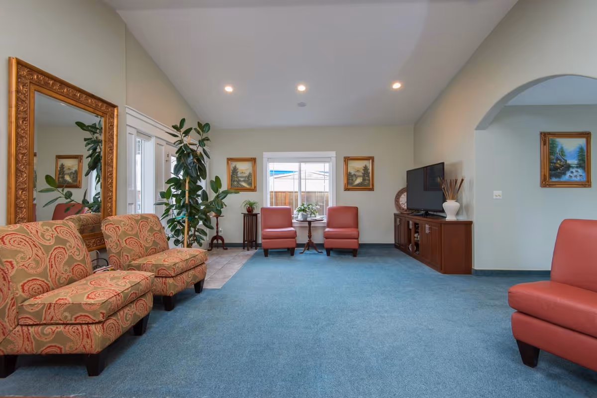 A spacious living room with blue carpet and beige walls. The room features two patterned armchairs on the left, two red armchairs near a window at the back, and another red armchair on the right. There is a large ornate gold-framed mirror on the left wall, several framed landscape paintings, a wooden TV cabinet with a flat-screen TV, and some potted plants adding greenery to the space.