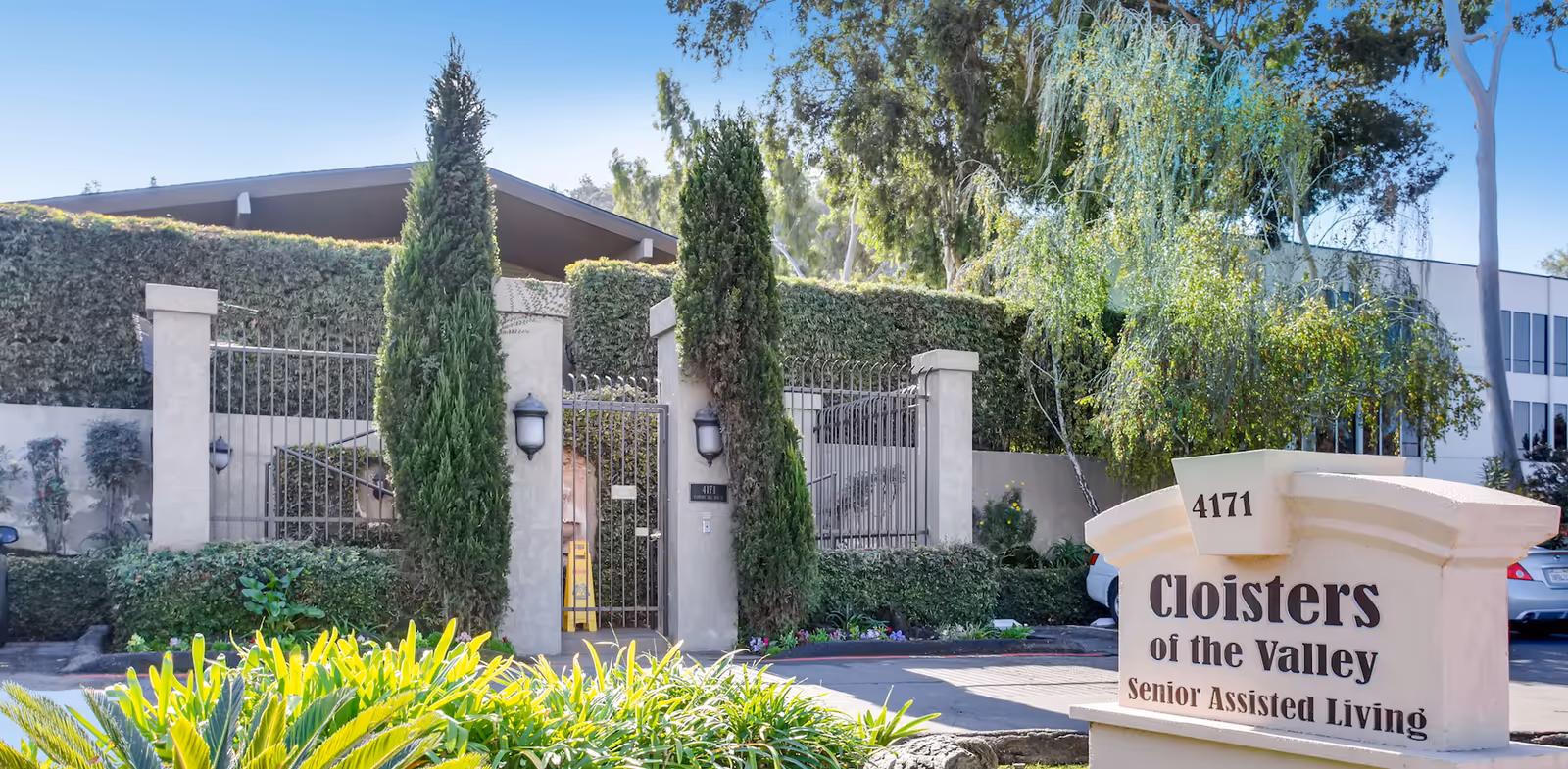 Exterior view of Cloisters of the Valley Senior Assisted Living facility showing a gated entrance with tall green hedges and trees, and a stone sign with the facility name and address 4171.