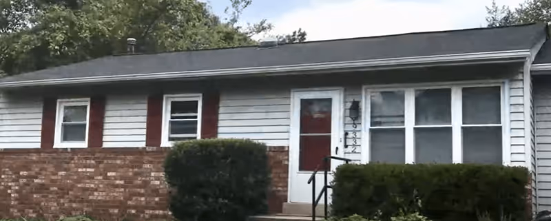 Exterior front view of a single-story residential building with white siding and red brick lower facade, featuring three windows with red shutters, a white door with a red panel, and trimmed bushes in front.
