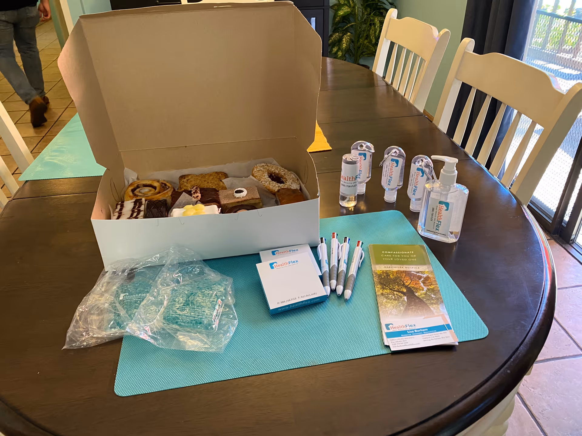 A dark wooden dining table with a light blue placemat displaying a box of assorted pastries, several pens, hand sanitizers, brochures, and some plastic packaging. White chairs surround the table and a person is walking away in the background on a tiled floor.