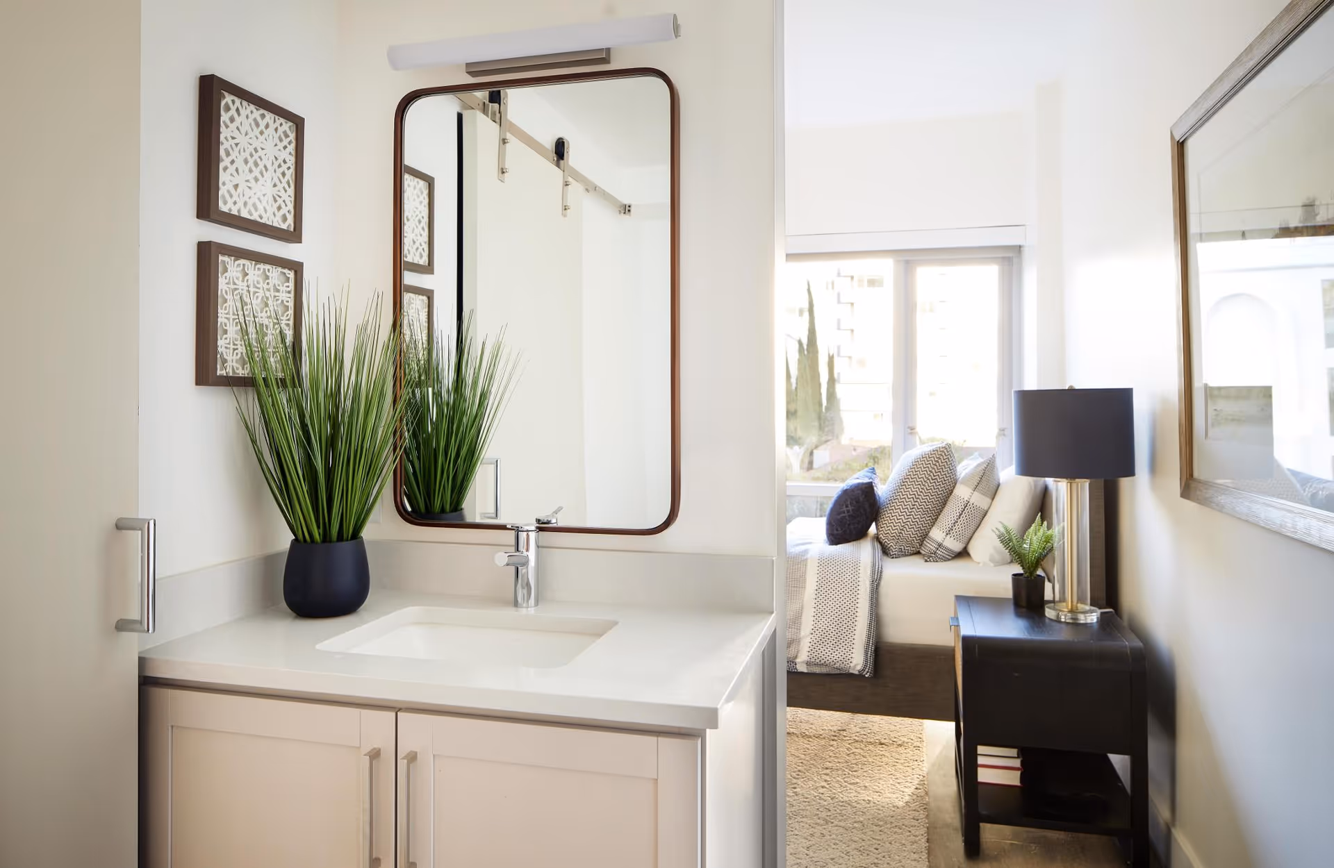 A bright and modern bathroom vanity area with a rectangular mirror above a white countertop sink. A black vase with tall green grass-like plants sits on the countertop. To the right, there is a view into a bedroom with a bed adorned with multiple pillows and a bedside table holding a lamp and a small plant. The room has a large window letting in natural light.