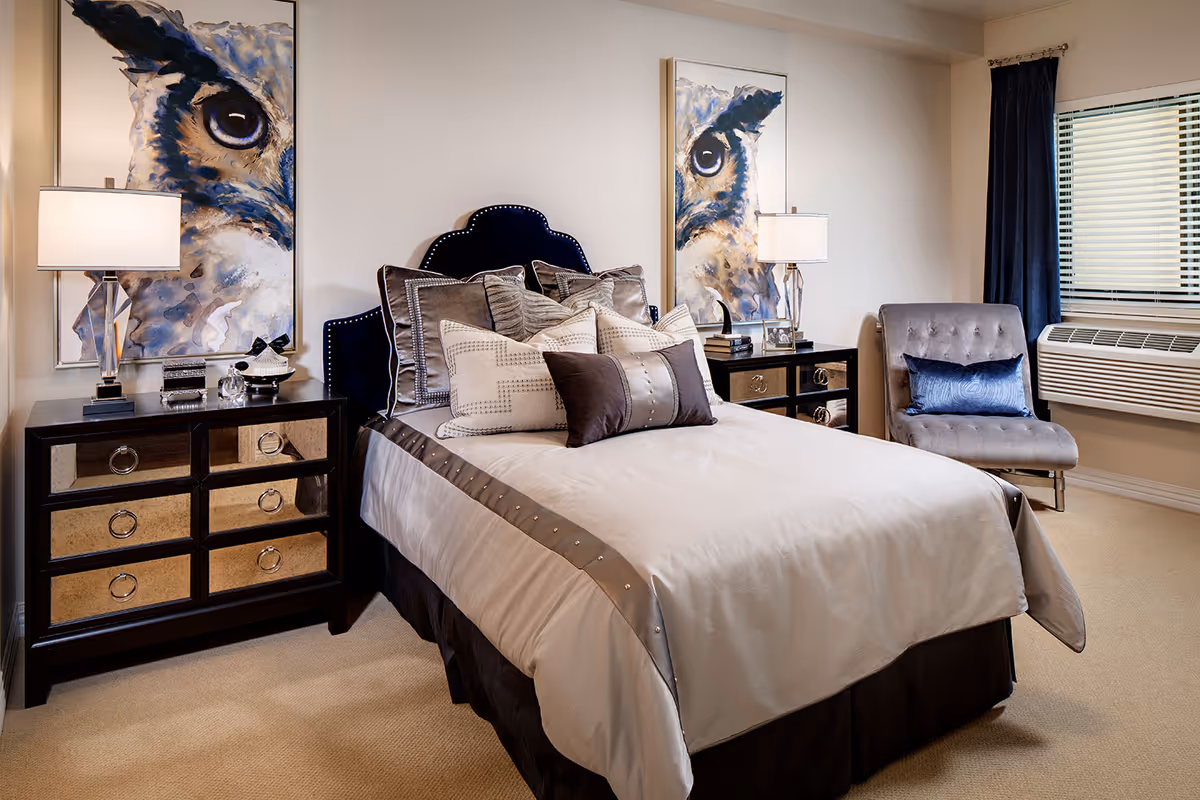 A neatly made bed with multiple decorative pillows in a senior living bedroom. The bed has a dark blue upholstered headboard with nailhead trim. On either side of the bed are matching dark wood nightstands with mirrored drawers, each topped with a modern table lamp and decorative items. Two large paintings of an owl's face hang on the wall behind the nightstands. A gray upholstered chair with a blue pillow is positioned near a window with blinds and dark curtains. The room has beige carpeting and neutral-colored walls.
