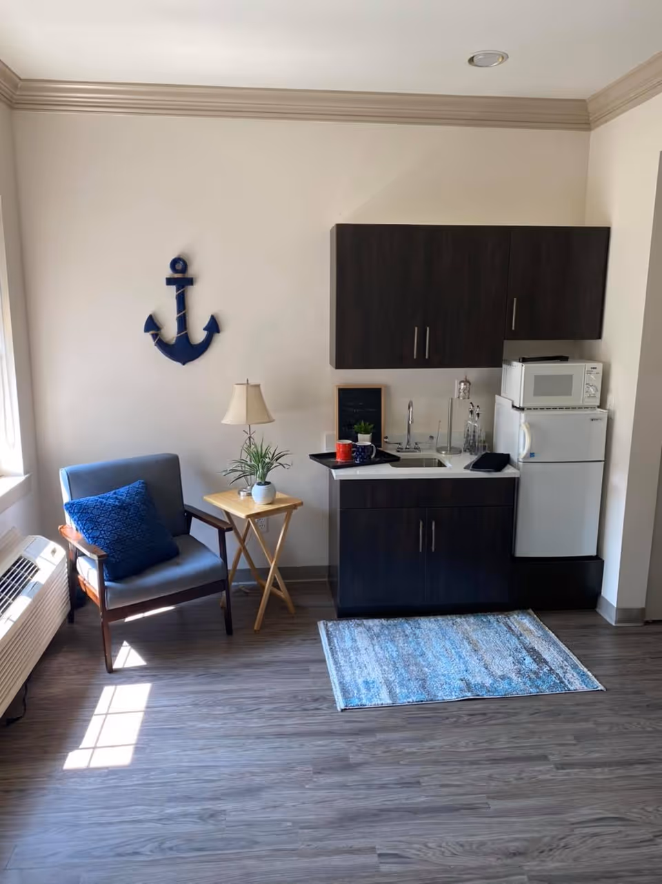 A small kitchenette area with dark wood cabinets, a white countertop with a sink, a microwave on top of a mini refrigerator, and a blue patterned rug on the floor. To the left, there is a gray armchair with a blue cushion, a small wooden side table with a lamp and a potted plant, and a blue anchor wall decoration. Sunlight streams in from a window on the left side.