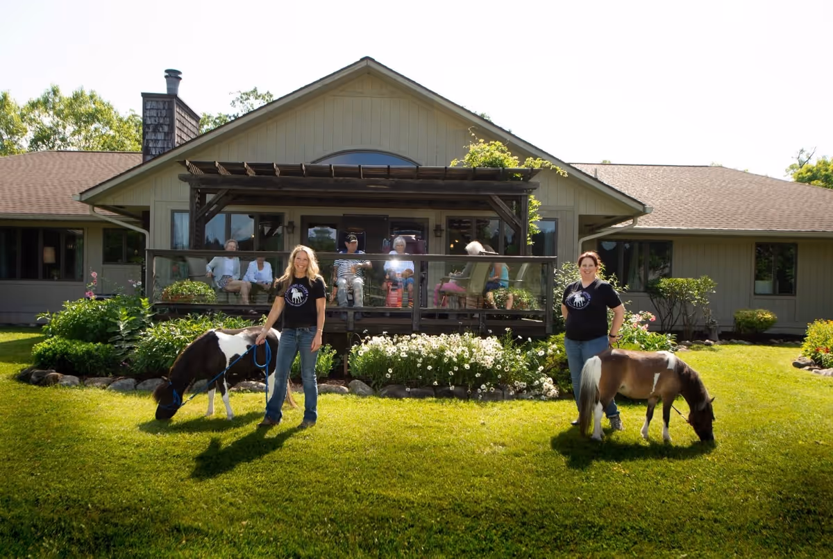 Front view of a single-story assisted living building with residents on a porch and two women holding small ponies on the lawn.